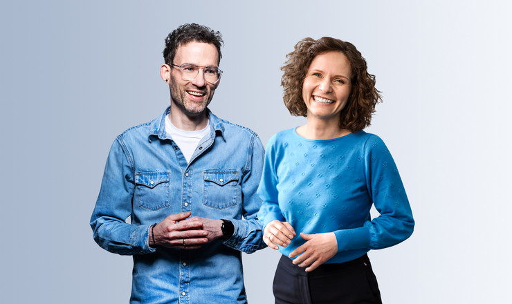 Zwei freundlich lächelnde Personen stehen vor hellem Hintergrund und blicken offen in die Kamera.